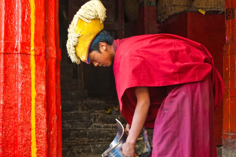 A monk in crimson robes pouring butter tea from a metal kettle beside a monastery doorway