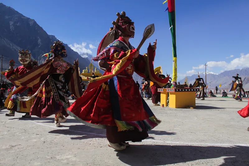 Masked dancers in brocade robes mid-turn on a monastery courtyard, mountains rising behind them