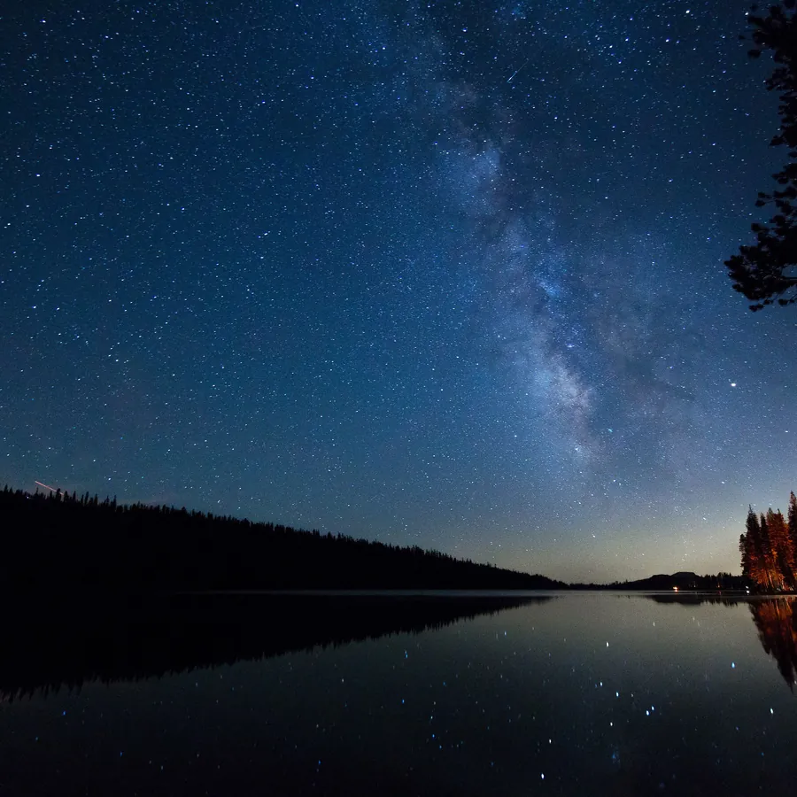 The Milky Way over a still high-altitude lake at night, the far shore a black ridge against the stars